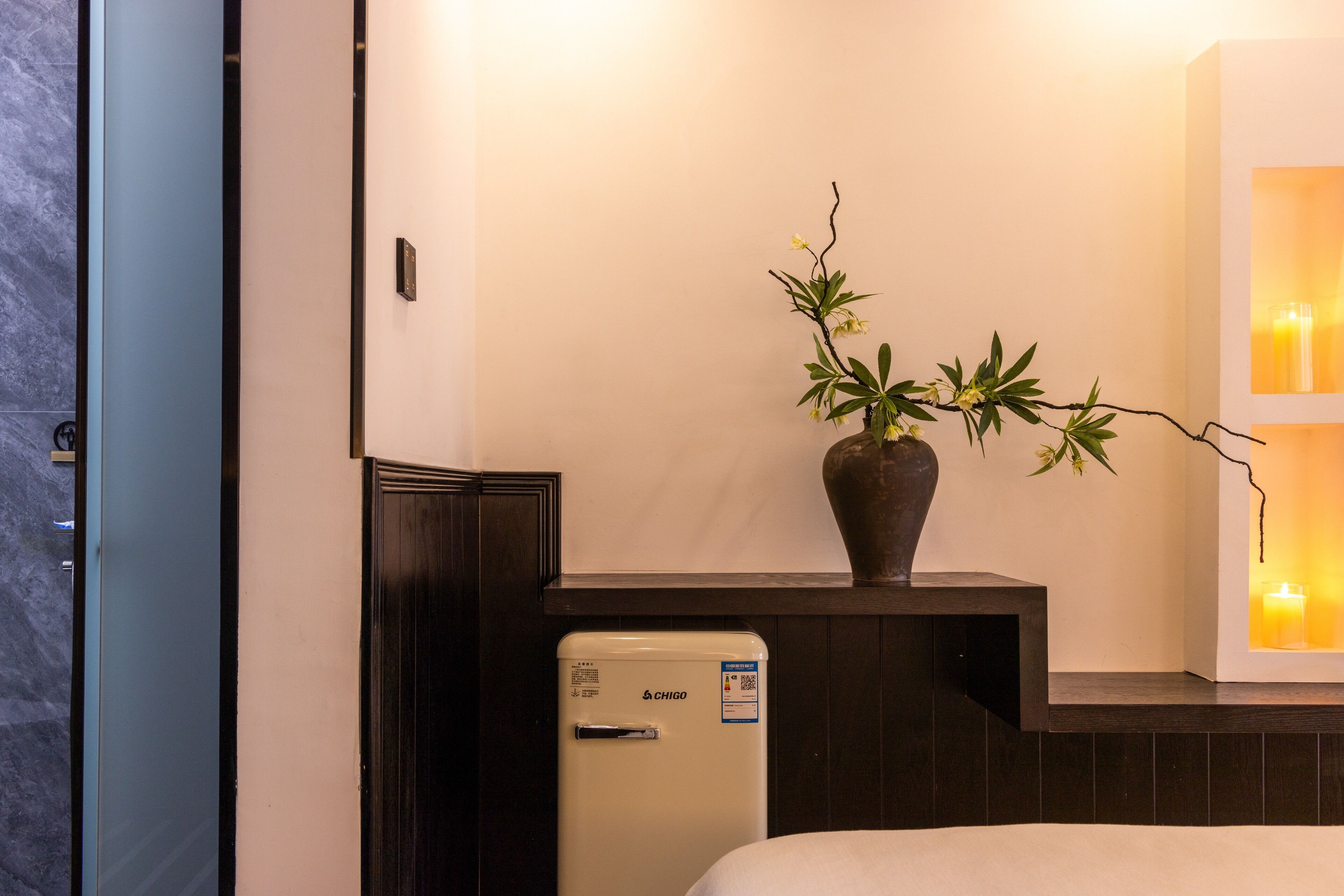 A compact room with a bed, a small white refrigerator, a vase with a plant, and a wall-mounted light fixture.