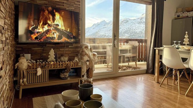 A cozy living room with a fireplace, a television displaying a fire, a dining area with a table and chairs, and a view of snow-capped mountains through large sliding doors. - Le Refuge du Loup - Panoramic view, Netflix & Pool (Dévoluy)