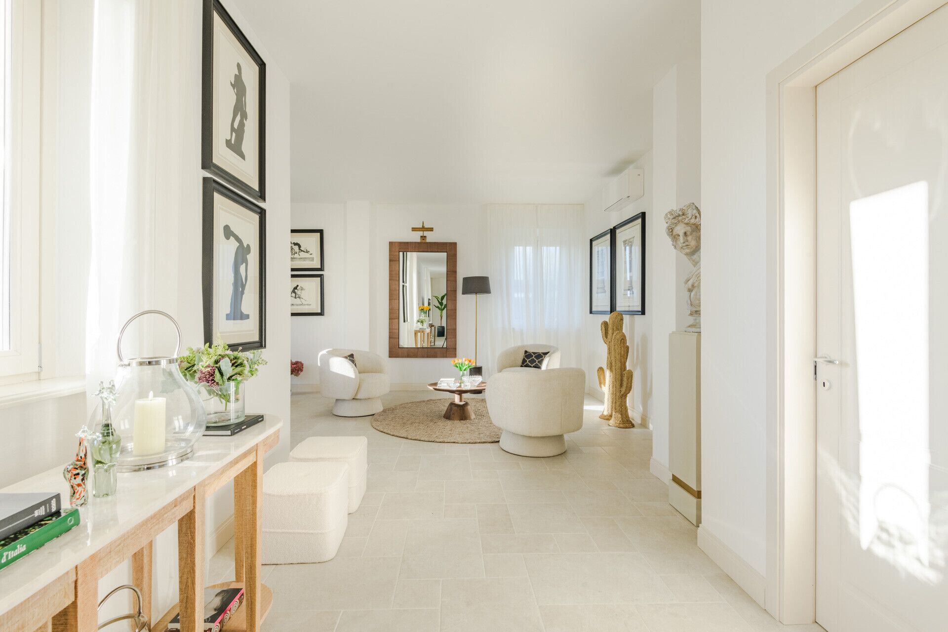 Un pasillo amplio con piso blanco, una consola de madera con un florero y libros, un sillón blanco, una mesa de centro redonda y un espejo.