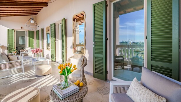 Una habitación iluminada por el sol con muebles de mimbre, contraventanas verdes y vistas al campo.