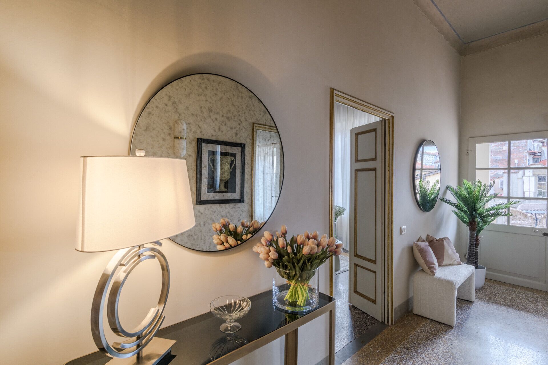 A hallway with a round mirror, a table with a lamp, a vase with flowers, and a potted plant.