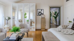 A modern living room with a sofa, armchair, and a coffee table with books. There is a large mirror and a door leading to another room. - The Marble Penthouse (Lucca)