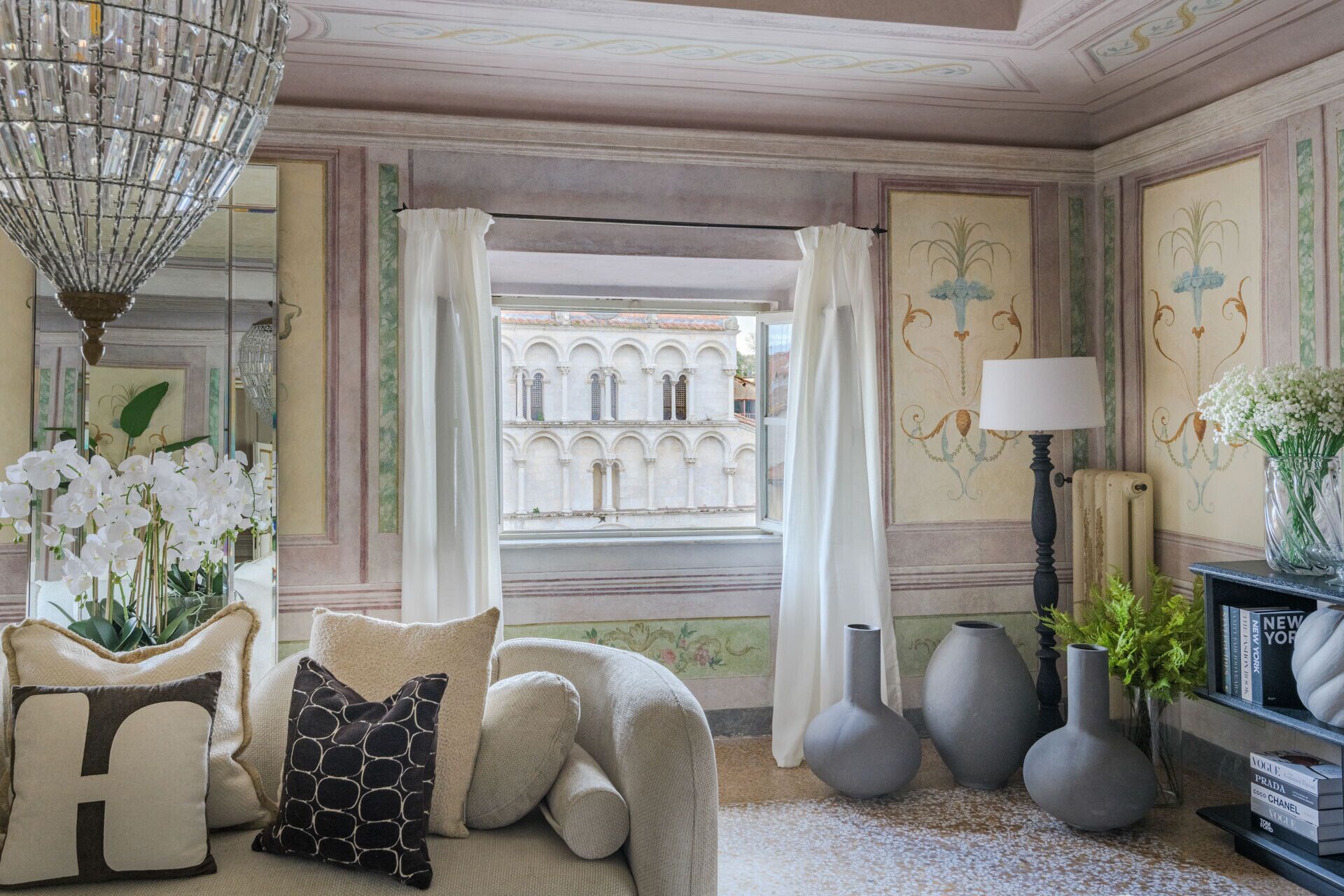A room with a view of a historic building, a chandelier, and a sofa with decorative pillows.