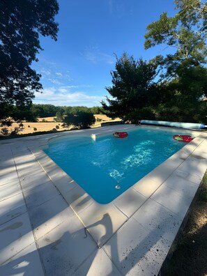 Ein rechteckiger Swimmingpool mit Fliesen umgeben von Bäumen und blauem Himmel.