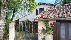 Uma casa de pedra rústica com telhado de cerâmica, um portão de metal e um pequeno jardim com flores vermelhas.