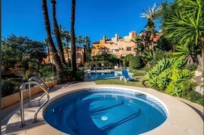 Une piscine dont le carrelage présente un motif géométrique, entourée de palmiers et de verdure, avec un bâtiment en arrière-plan.