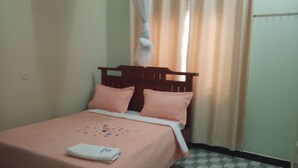A single bed with a wooden headboard, a pink bedspread, and matching pillows. A white towel is folded on the bed. The room has a checkered floor pattern and beige walls.