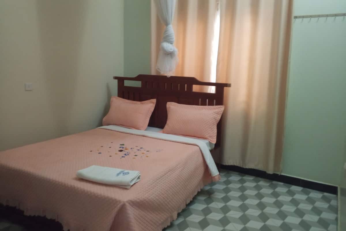 Een eenpersoonsbed met een houten hoofdbord, een roze dekbedovertrek en passende kussens. Een witte handdoek ligt op het bed gevouwen. De kamer heeft een geruit vloertapijt en beige wanden.