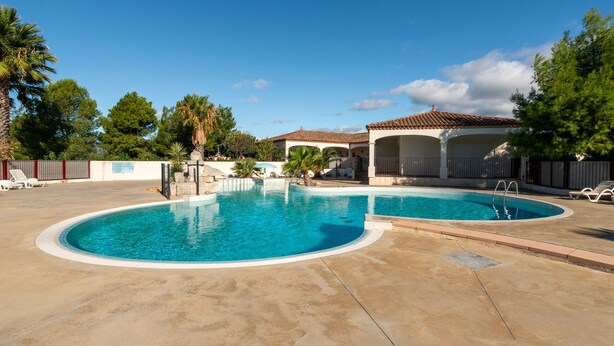 A swimming pool with a curved design, surrounded by a concrete deck. There are lounge chairs and a covered area with a tiled roof. - Charming villa with garden for 6 pers (Homps)
