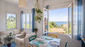 Habitación con sofá, mesa de comedor con individuales, una planta colgante y vista al mar.