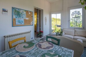 Ein Zimmer mit Tisch, Stühlen, einer Couch, einem Fenster mit Baumdeko und einem Korkbrett mit Karten und Notizen.