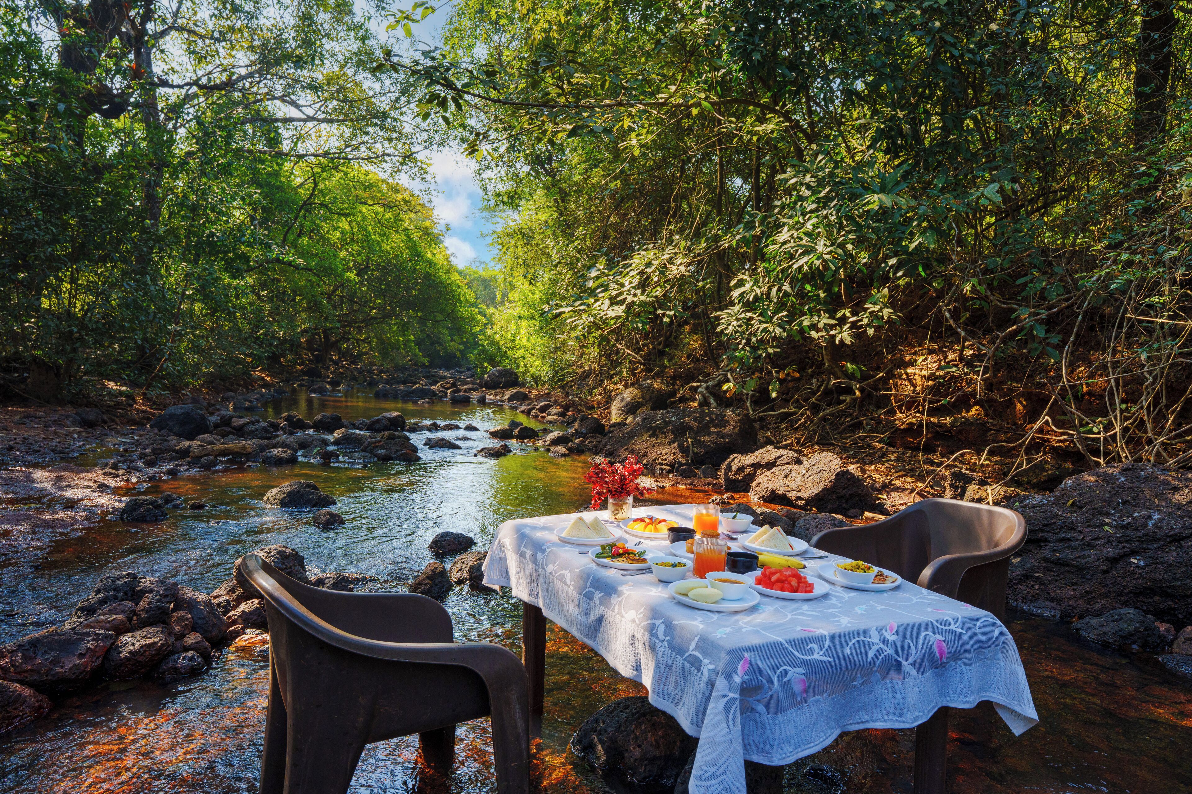Une table à manger installée au bord d’une rivière, avec une nappe et des assiettes remplies de nourriture.