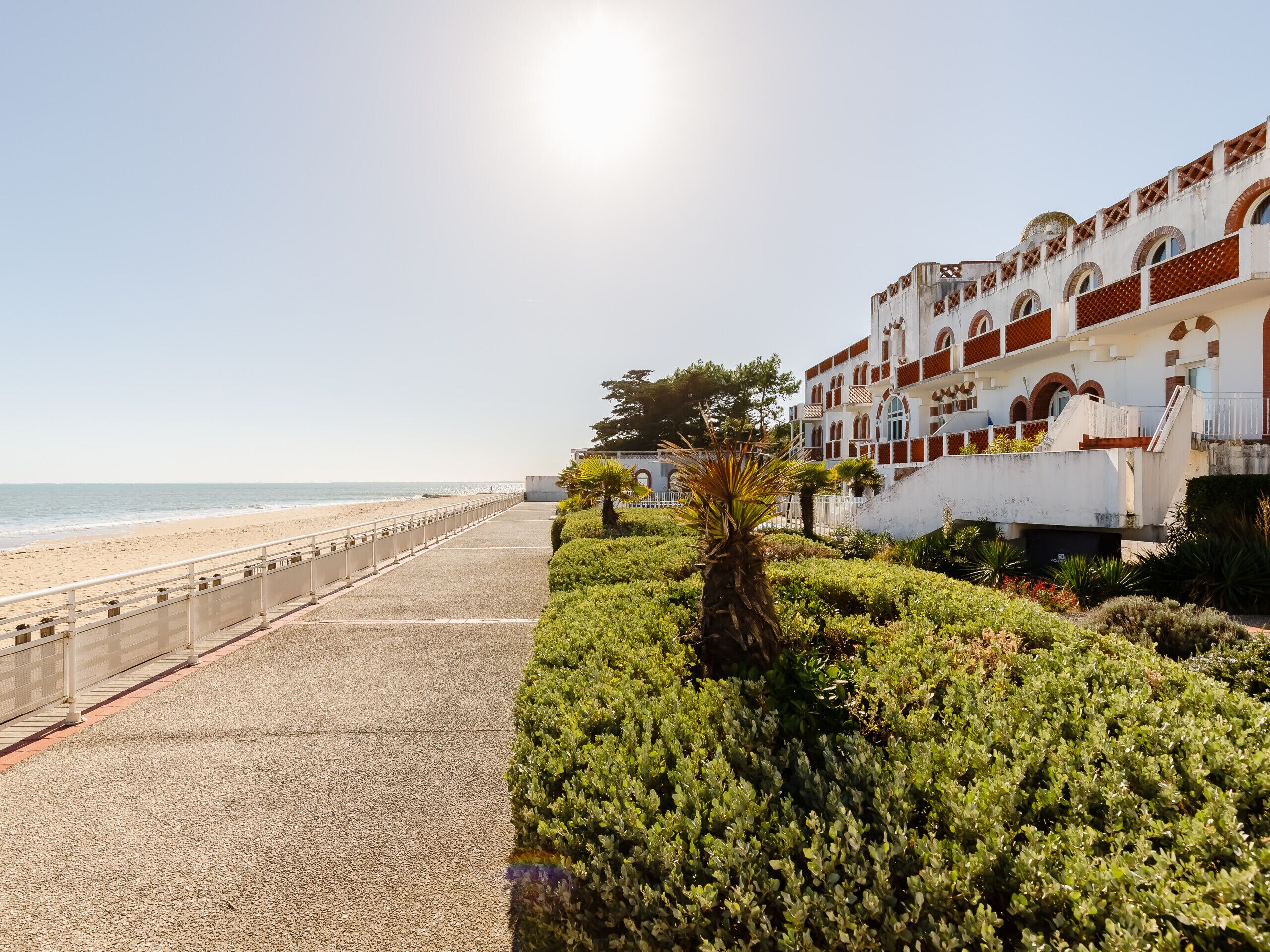 A coastal walkway with a beach view, a white building with red accents, and lush greenery.
