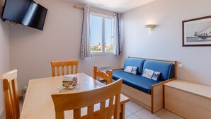 A room with a wooden table, chairs, a sofa with blue cushions, a television mounted on the wall, a window with curtains, and a framed picture on the wall. - Nice Apt 5 pers near the beach with pool (La Tranche-sur-Mer)