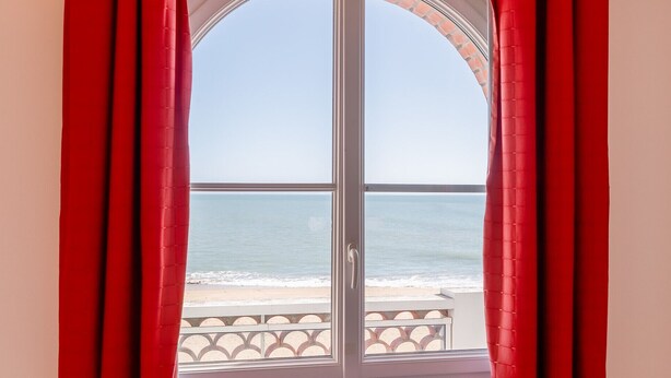 Ein Fenster mit roten Vorhängen, Blick auf den Ozean und einem Balkongeländer.