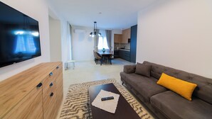 Een moderne woonkamer met een flatscreen televisie, een bruine bank met een geel kussen, een houten kast en een eethoek op de achtergrond.