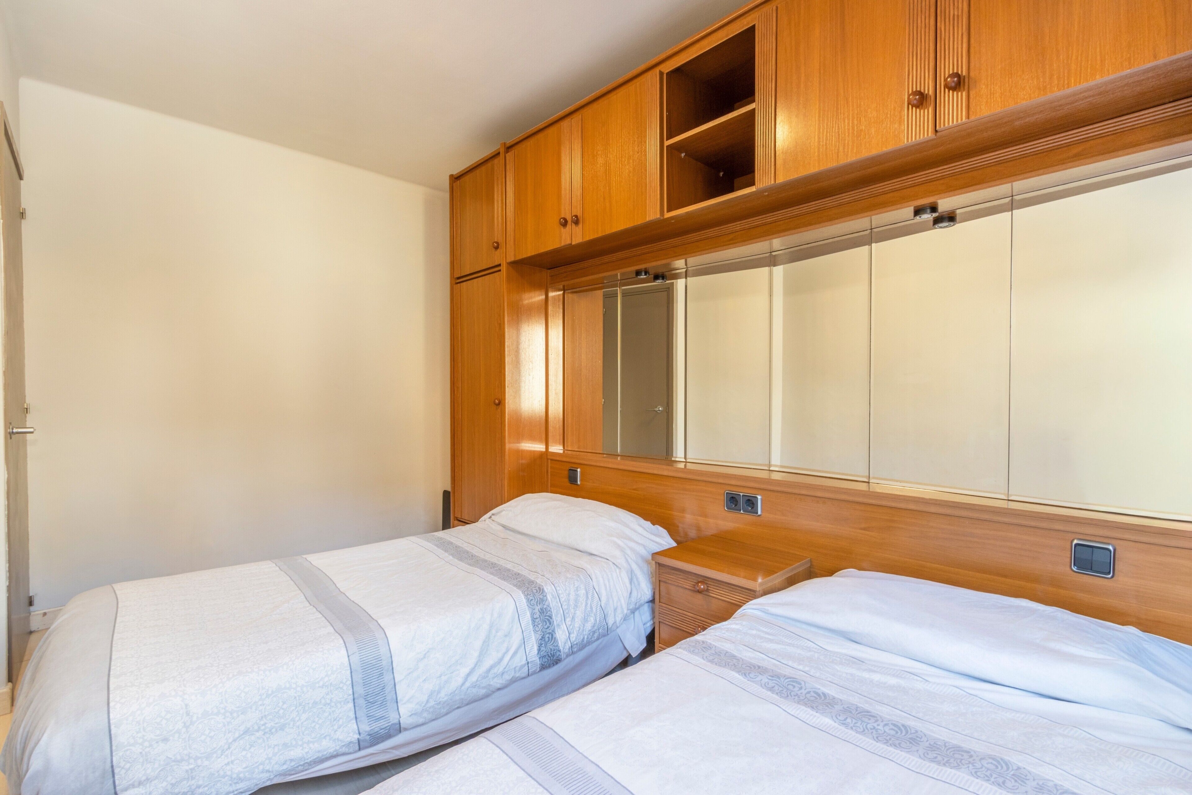 A bedroom with two beds, wooden cabinets, a large mirror, and a small bedside table.