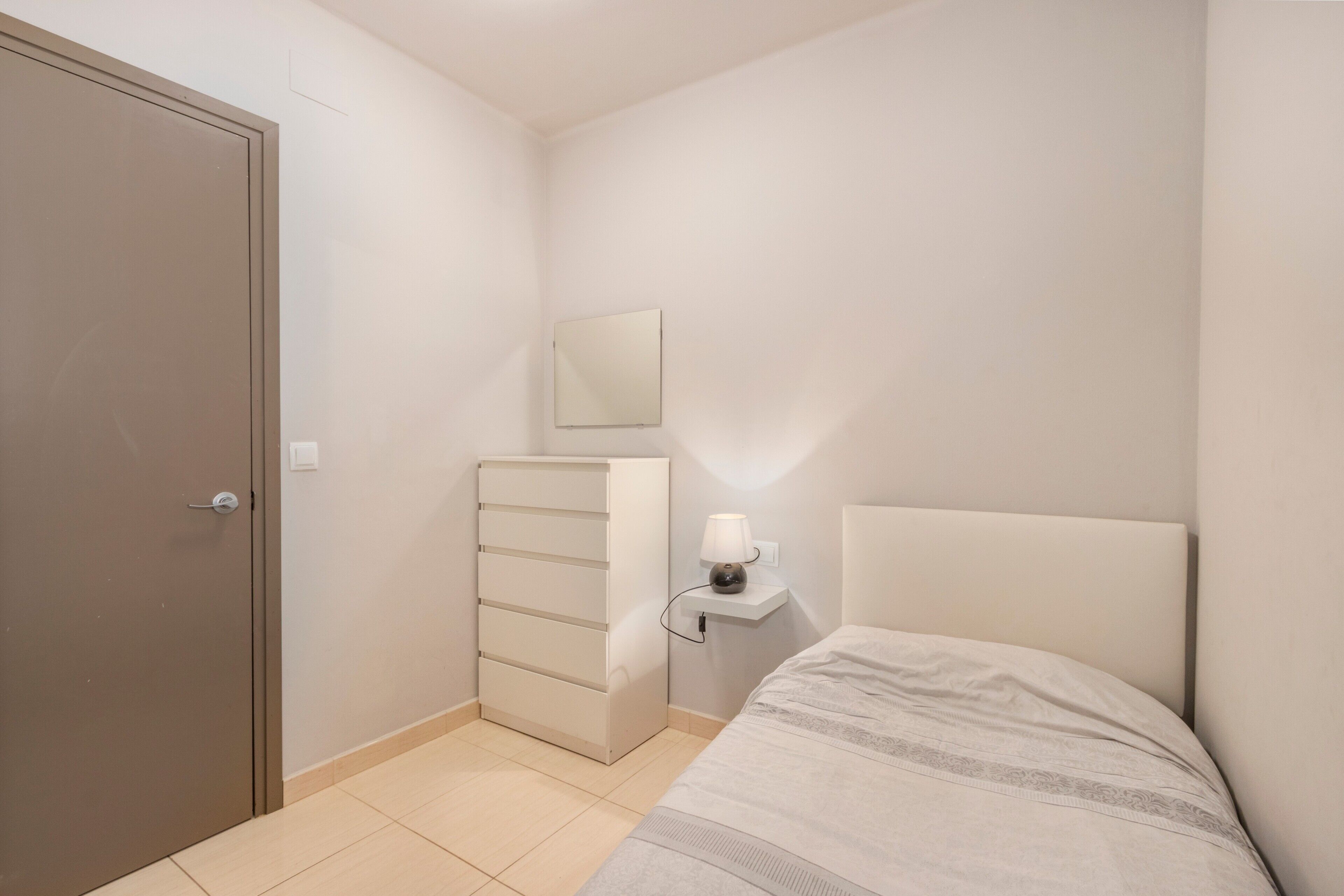 Ein Schlafzimmer mit Bett, Kommode, Nachttisch mit Lampe, Spiegel und Tür.