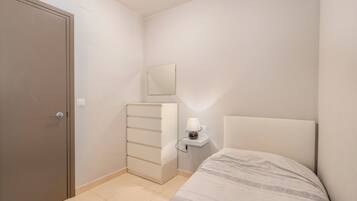 Ein Schlafzimmer mit Bett, Kommode, Nachttisch mit Lampe, Spiegel und Tür.