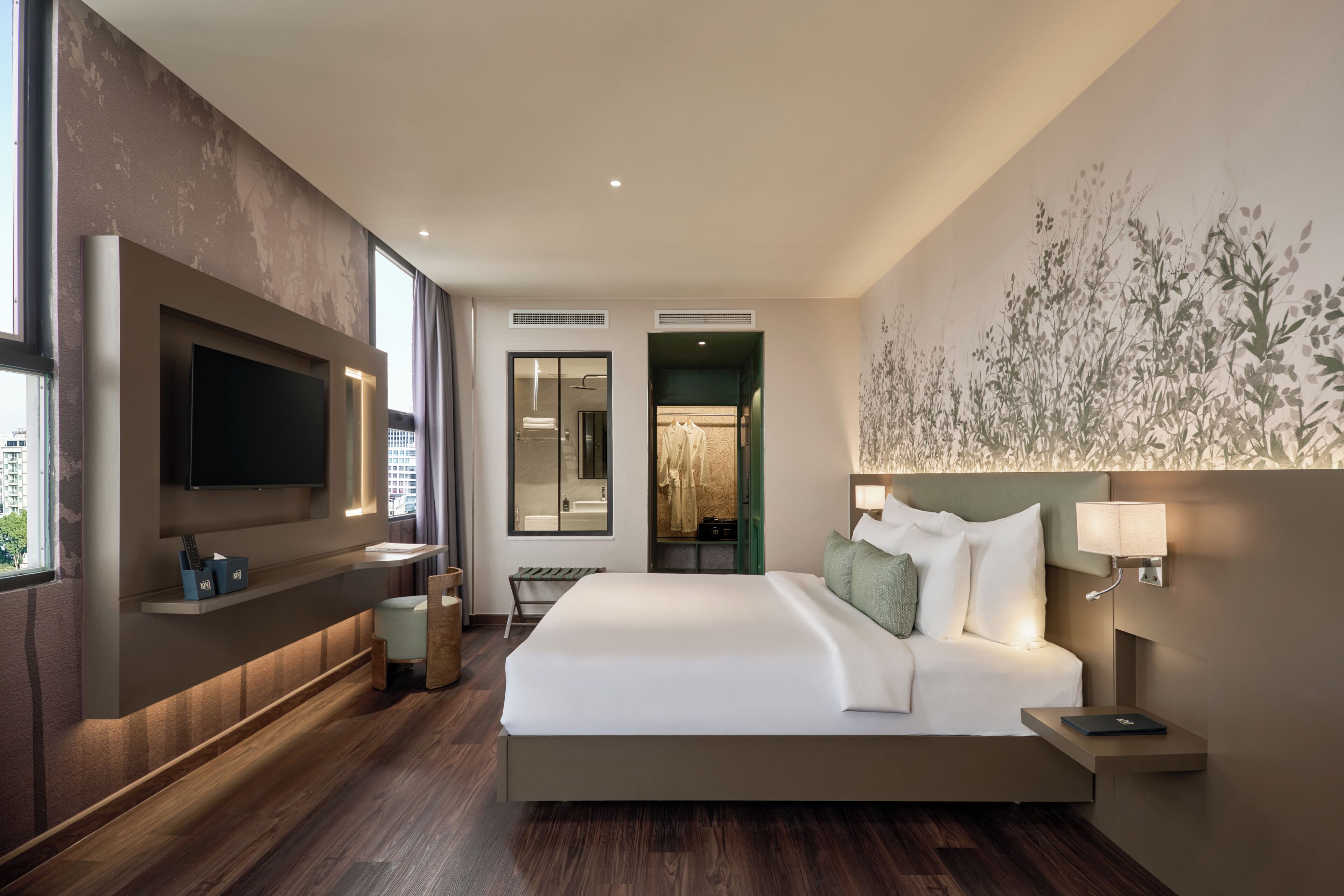 Ein modernes Hotelzimmer mit einem großen Bett, einem flachbildfernseher an der Wand, einem Nachttisch mit Lampe und Blick auf die Stadt durch das Fenster.