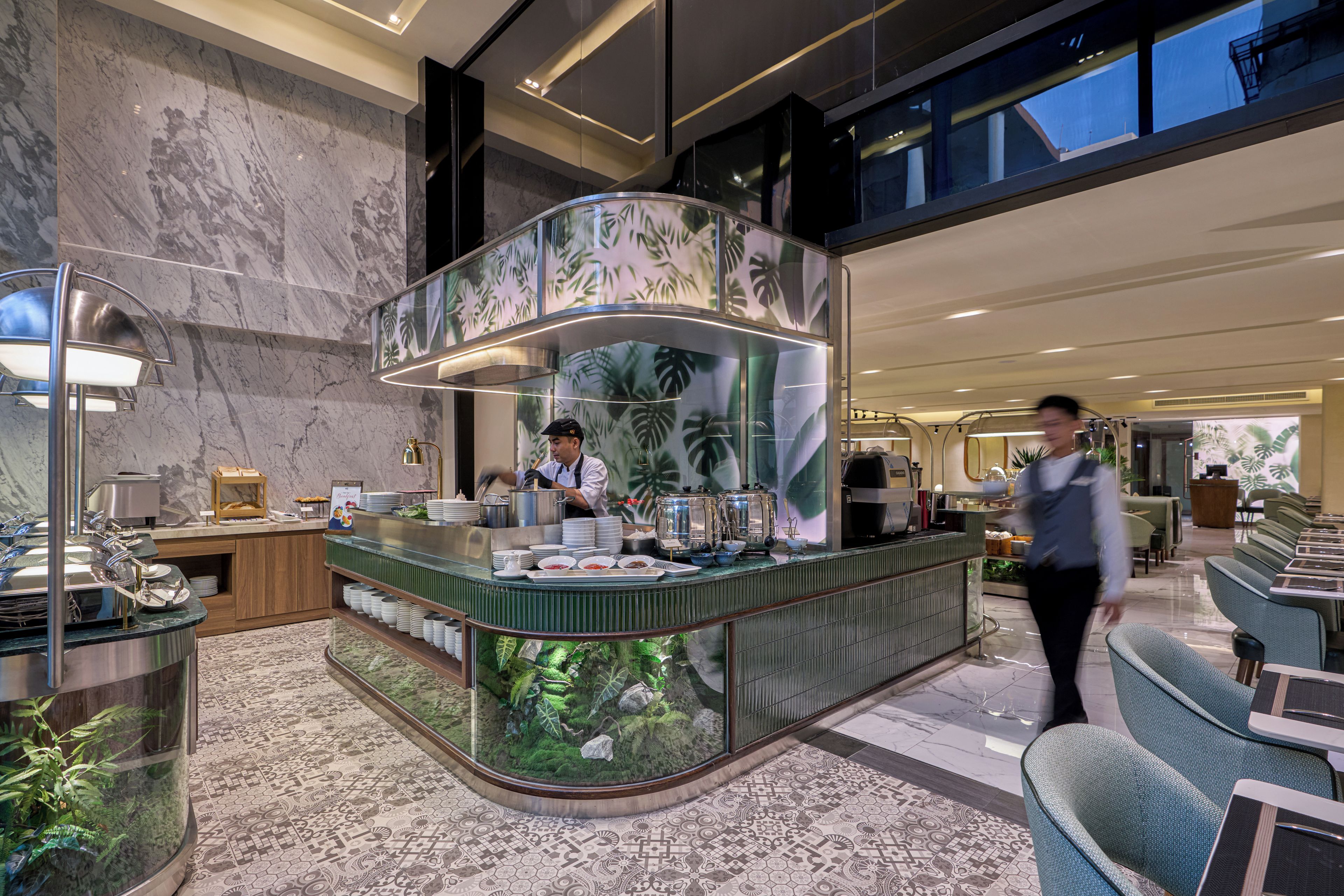 A hotel lobby with a marble floor, a buffet counter with a tropical leaf design, and seating area with green upholstered chairs.