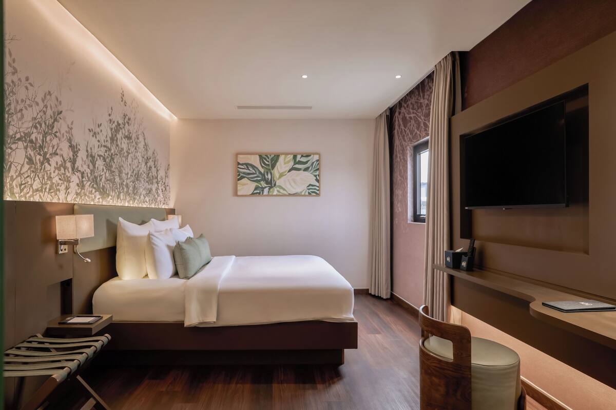 A modern hotel room with a large bed, a flat-screen TV, a desk with a laptop, and a wall-mounted artwork.