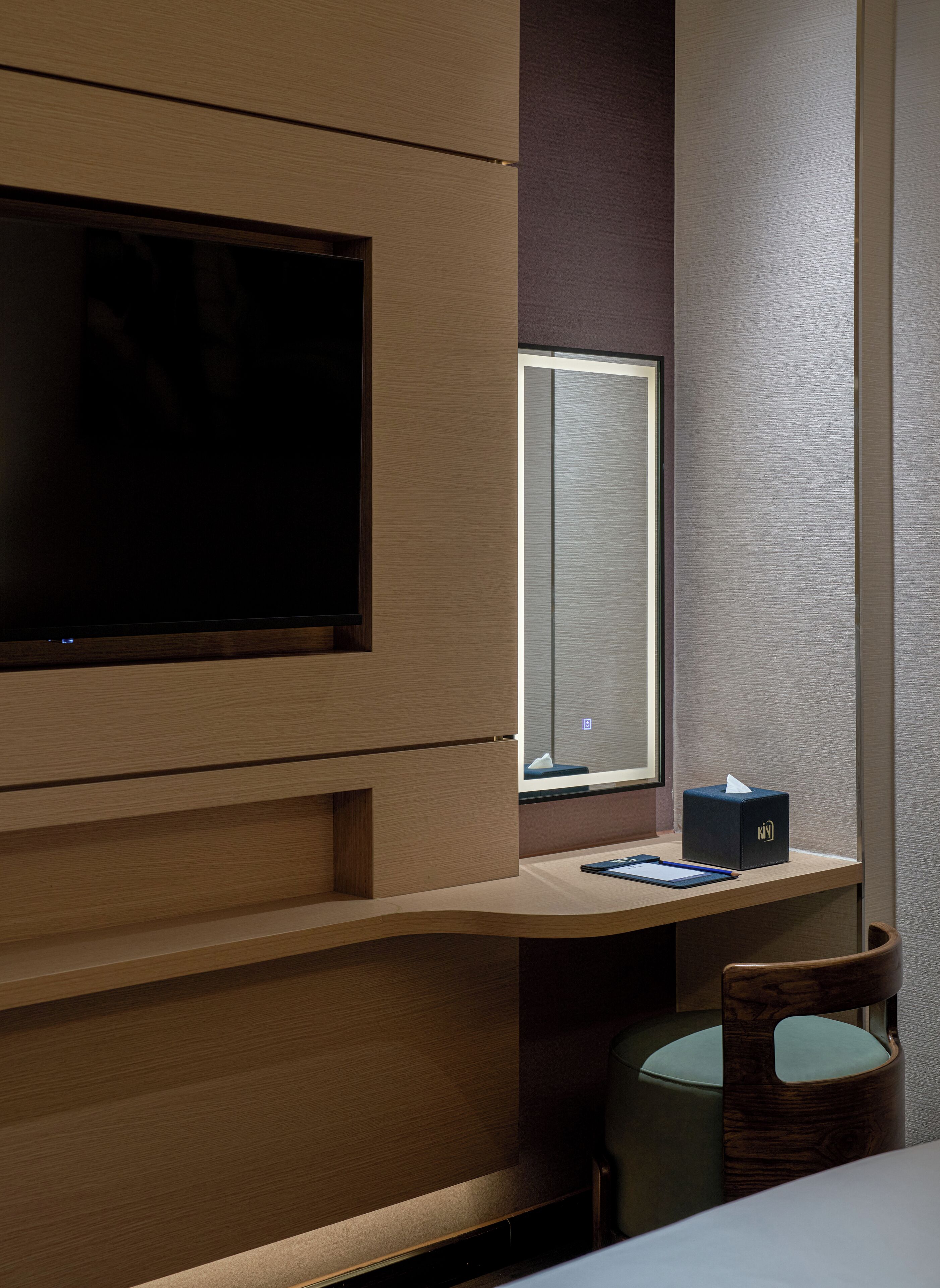Habitación de hotel moderna con un televisor de pantalla plana grande montado en un panel de madera, una ventana pequeña con vistas, un escritorio con una caja de pañuelos y una libreta, y una silla acolchada verde.