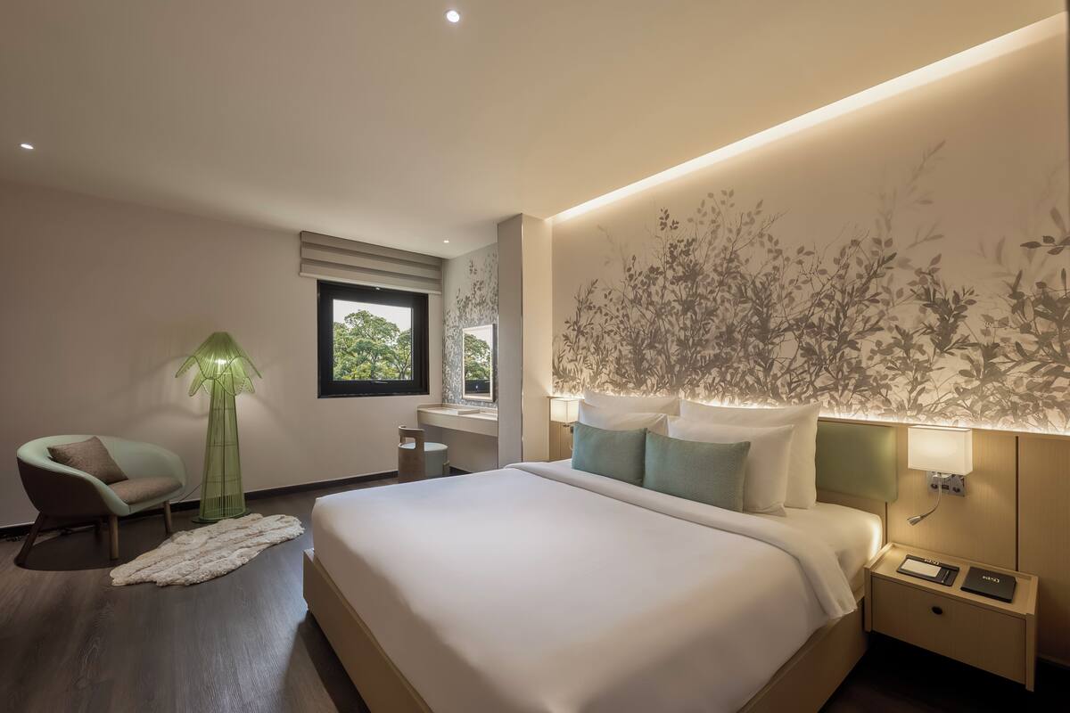 A modern hotel room with a large bed, bedside tables, a lamp, a chair, and a window.
