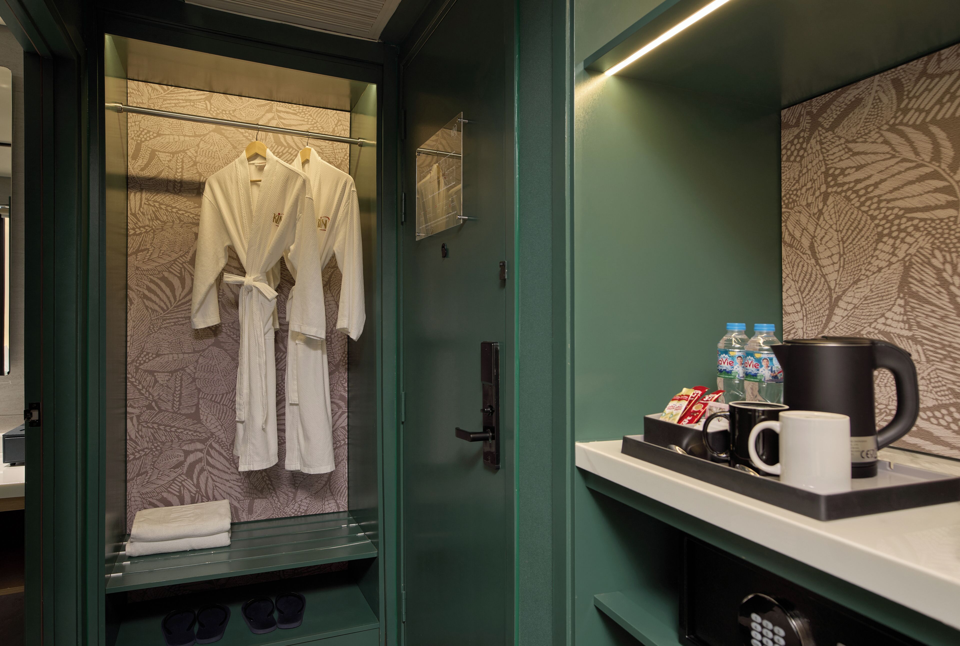 Habitación de hotel con puerta de vidrio para la ducha, perchero con batas, cafetera, agua embotellada y microondas sobre una encimera.