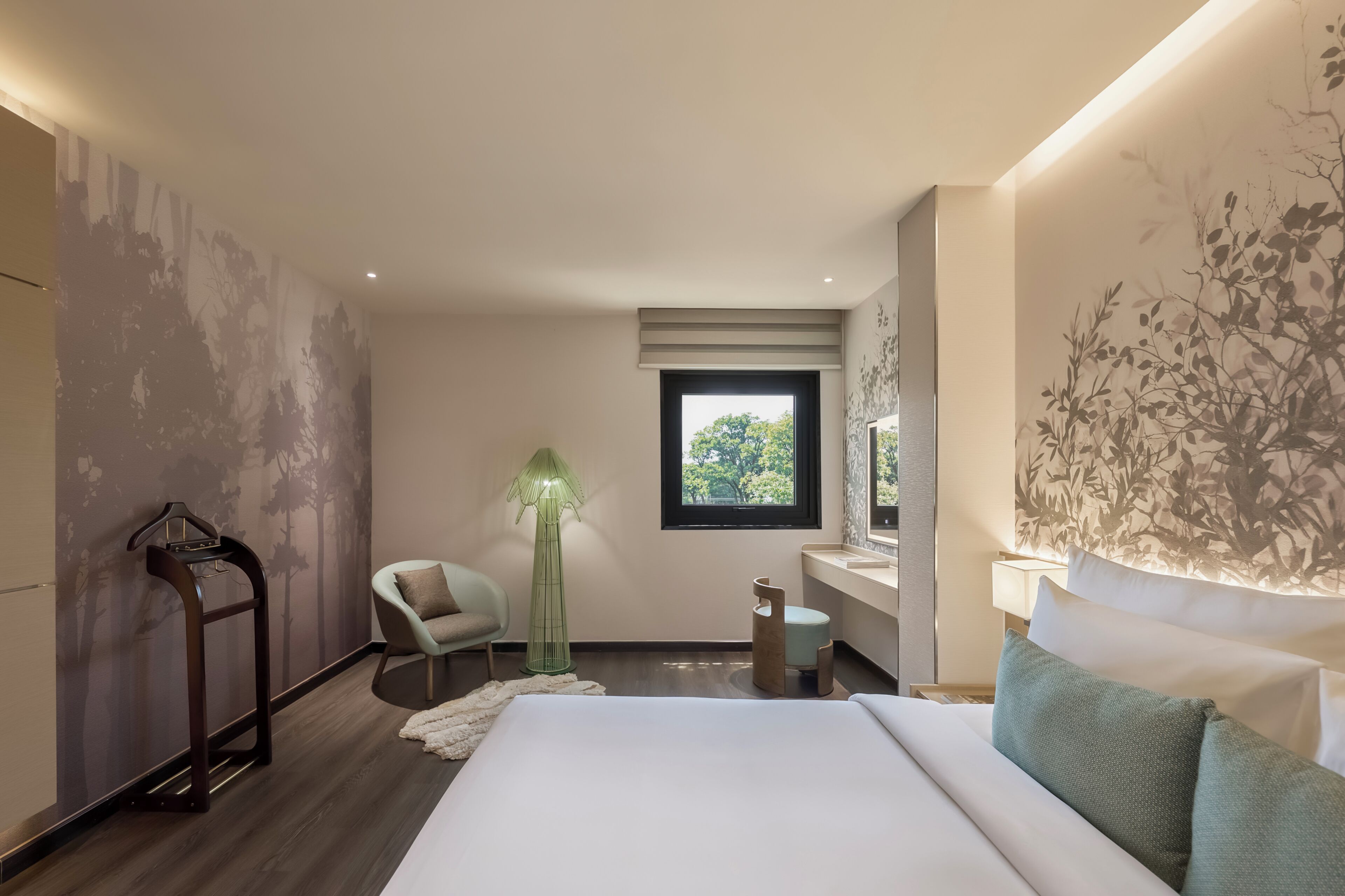 A modern hotel room with a large bed, a sitting area with a chair, a standing lamp, a window with a view, and a wall-mounted shelf.