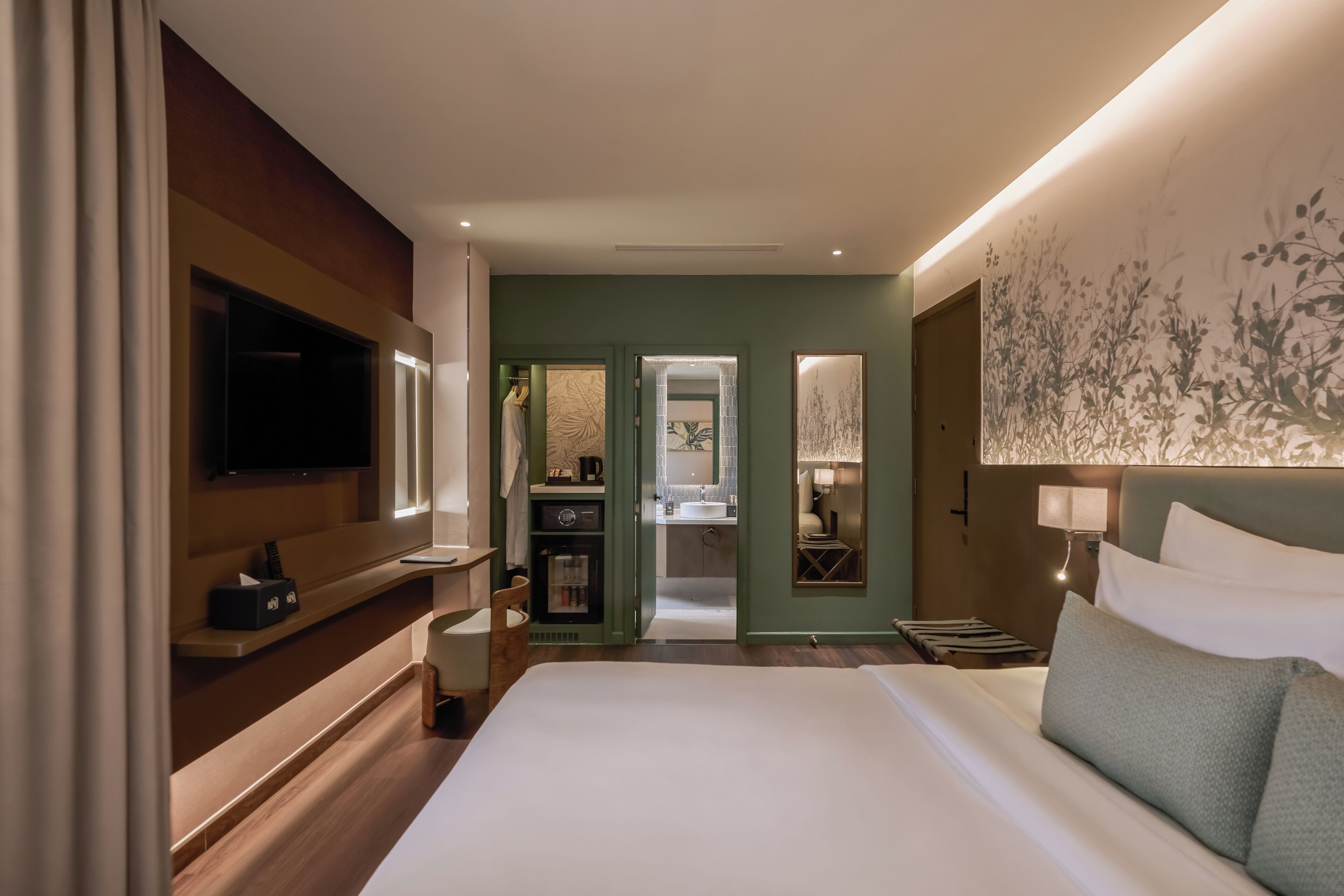 Ein Hotelzimmer mit einem großen Bett, einem an der Wand befestigten Fernseher, einem Schreibtisch mit Stuhl und einem durchgehenden Türspalte einsehbaren Badezimmer.