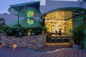 Un área de bar al aire libre con pared de piedra, terraza de madera y plantas tropicales.