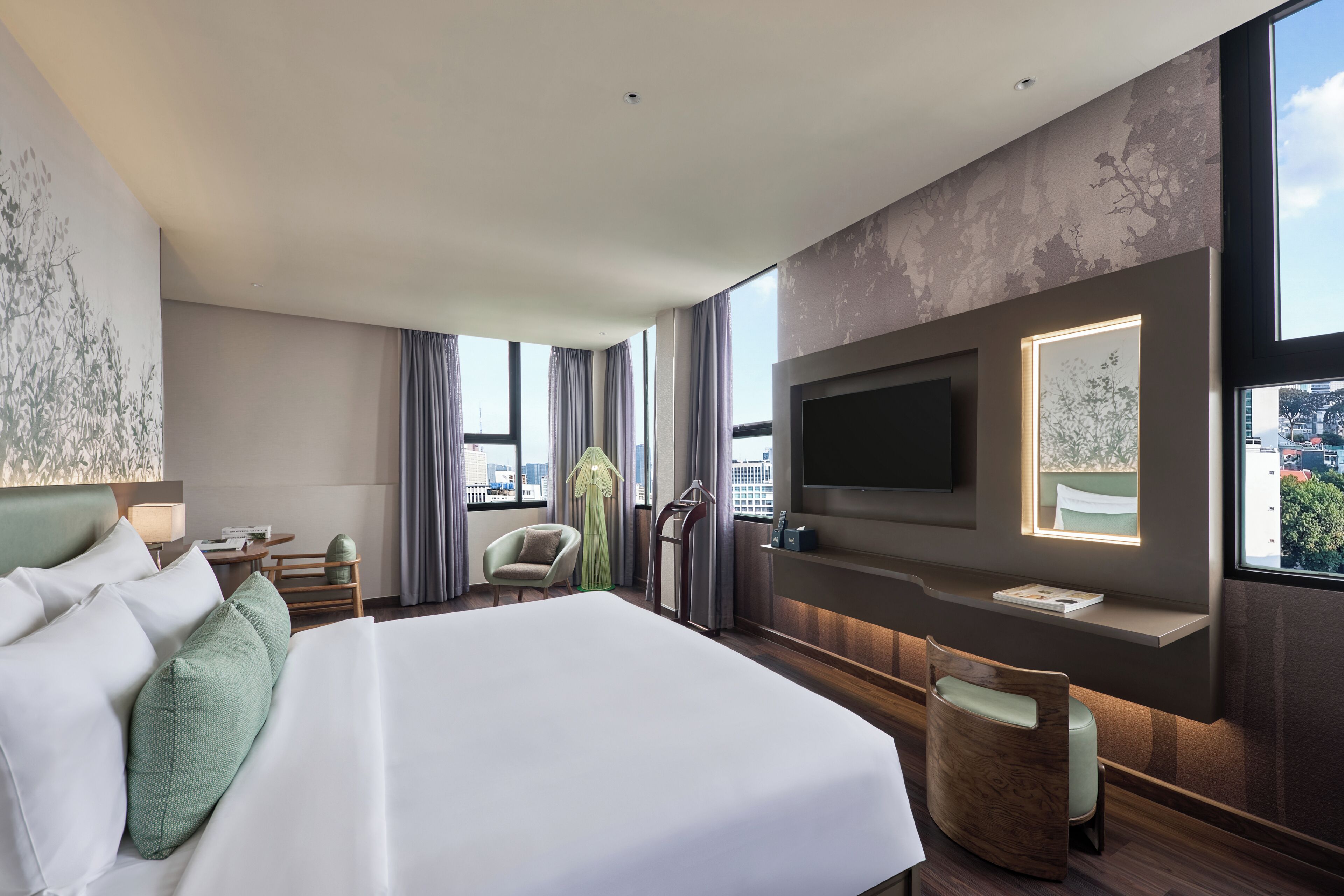 Quarto de hotel moderno com uma cama grande, televisão de tela plana na parede, área de estar com uma poltrona e vista para a cidade através de amplas janelas.