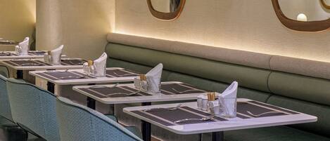 A restaurant interior with multiple tables and chairs, a green cushioned bench, and wall-mounted mirrors.