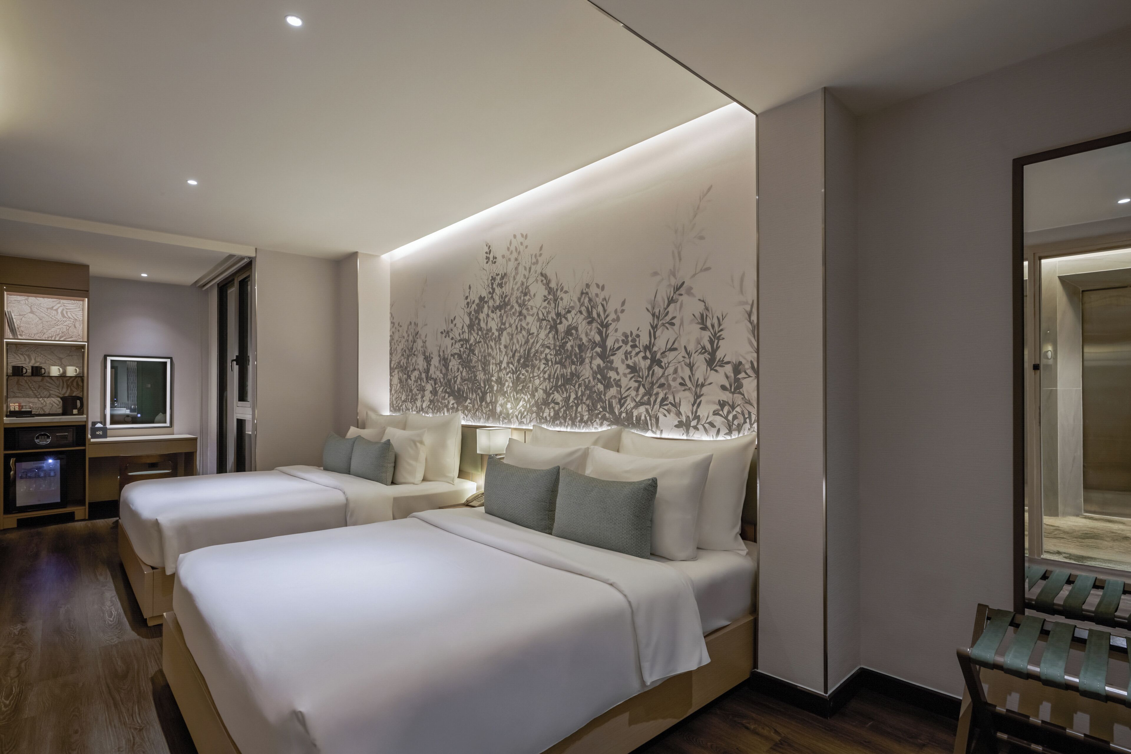 A hotel room with two beds, a desk, a minibar, and a large window.
