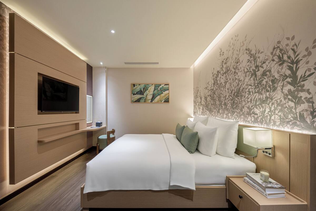 A modern hotel room with a large bed, a flat-screen TV mounted on the wall, a bedside table with a lamp, and a decorative plant wall.