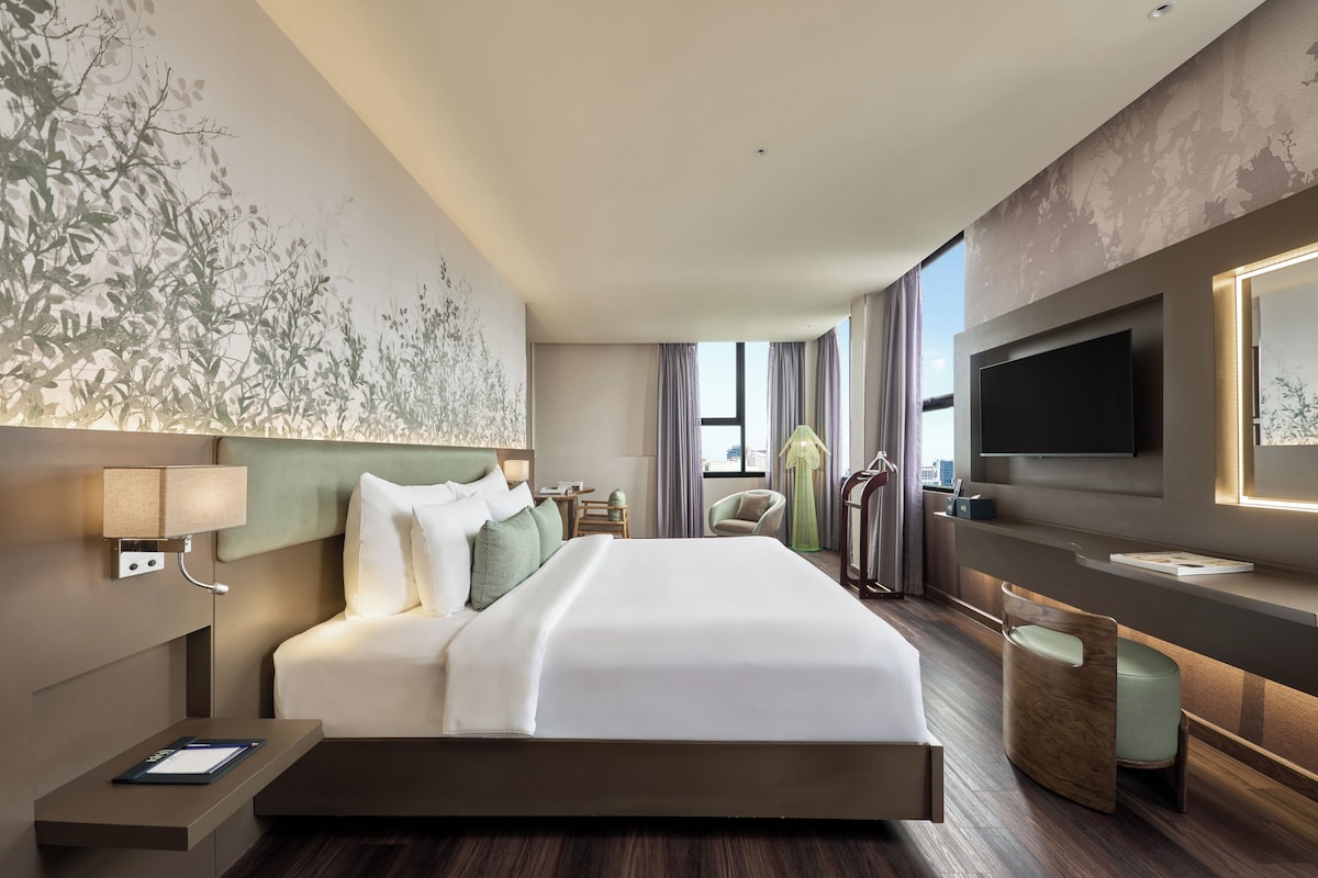 Habitación de hotel con una cama grande, un televisor en la pared, un escritorio con lámpara y vistas a la ciudad desde las ventanas.