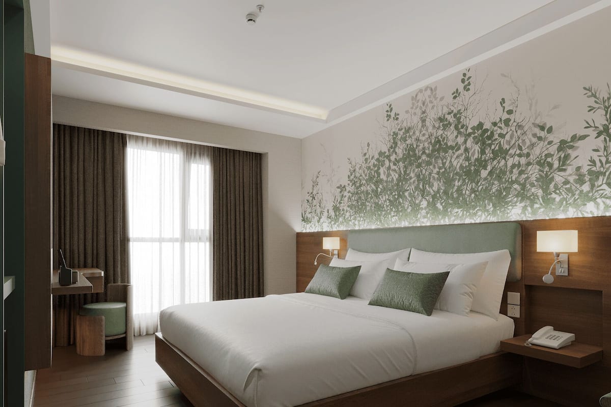 A modern hotel room with a large bed, a desk, and a chair. There is a window with curtains, a wall-mounted lamp, and a telephone on a bedside table.