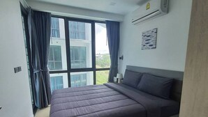 A modern bedroom with a large bed, a window with curtains, an air conditioning unit, and a small table with a lamp. - Stylish Bang Saray Apartment With Pool and Resort Facilities (Sattahip)