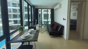 A modern living room with a dining area, large windows, and a view of a building. - Stylish Bang Saray Apartment With Pool and Resort Facilities (Sattahip)