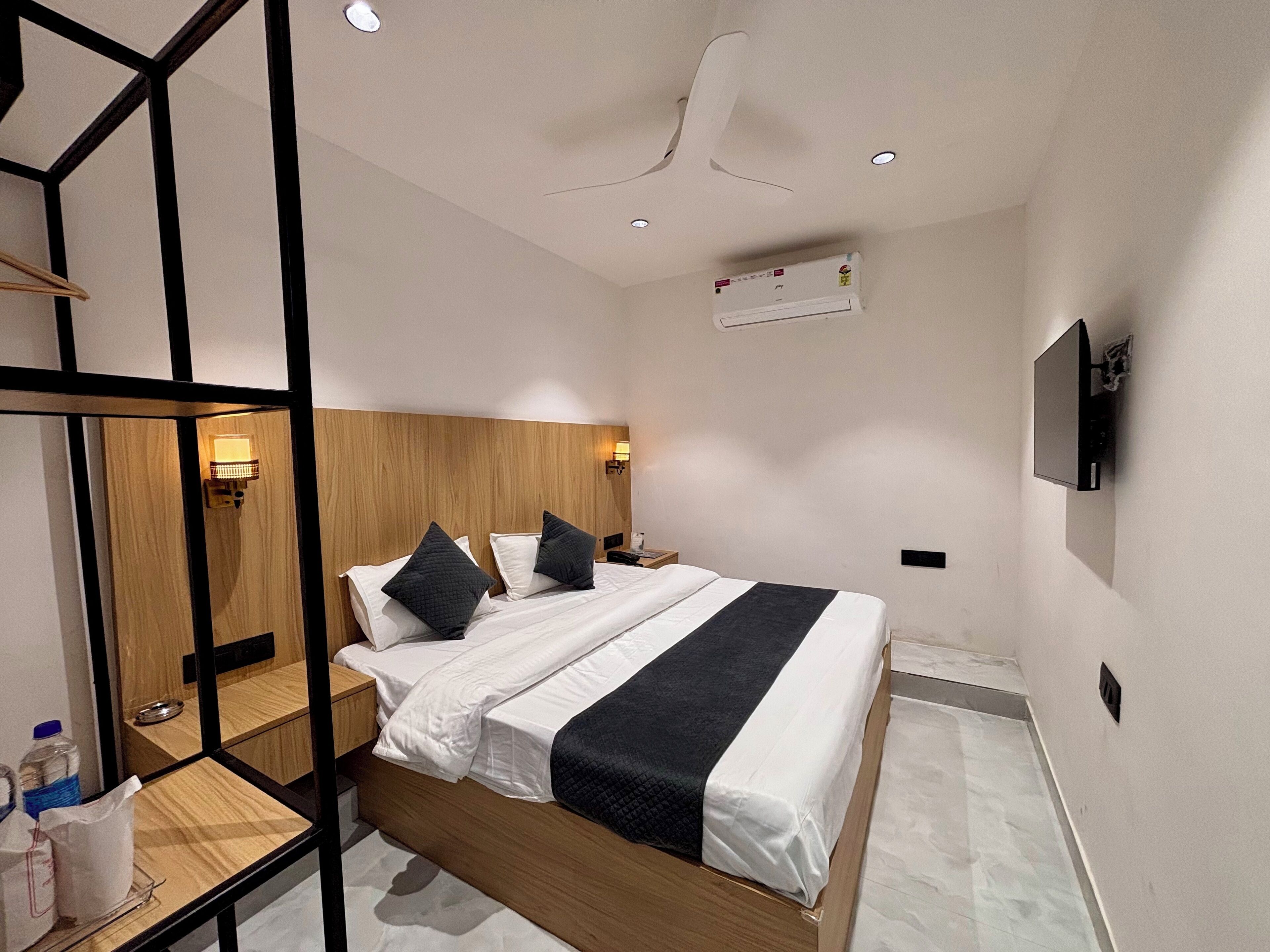 Una habitación de hotel moderna con cabecera de madera, una cama grande con ropa de cama blanca y negra, un televisor de pantalla plana montado en la pared y un ventilador de techo.