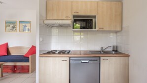 A compact kitchen with wooden cabinets, a microwave, a dishwasher, and a sink. There is a bed with red pillows and a small table in the adjacent room. - Cottage for 6 pers in a residence with pool (Ondres)