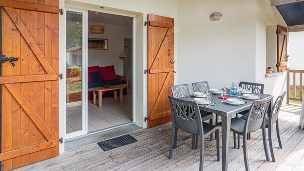 Una terraza de madera con una mesa lista para comer, un sofá con cojines rojos y una puerta que da a un área cubierta.