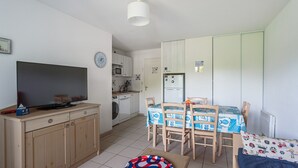 Un ambiente compatto con TV su un mobile in legno, un tavolo da pranzo con sedie, angolo cottura con frigorifero e lavatrice.
