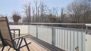 Ett trädäck med ett metallräcke, en stol och utsikt över lövträden under en klar himmel.