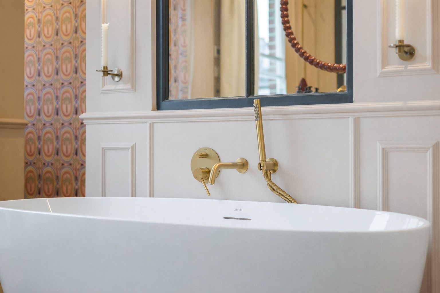 A white freestanding bathtub with gold fixtures in a bathroom with patterned wallpaper and a mirror above it.