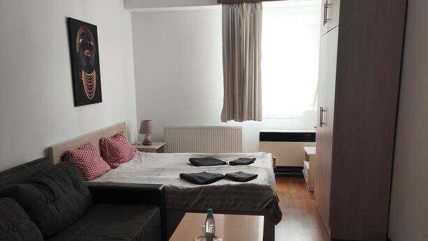 A modern hotel room with a dark-colored sofa, a small wooden table with a bottle and glasses, a bed with a white duvet, and a window with curtains. - Snowy Loft Didveli (Bakuriani)
