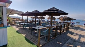 Un restaurant a la vora de la platja amb seients a l'aire lliure, veles de palla i vista al mar.