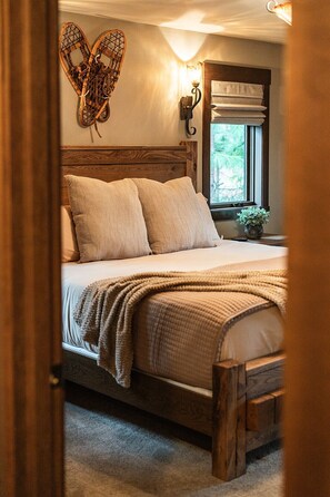 Une chambre avec un lit en bois, une literie de couleur beige, un luminaire fixé au mur et une fenêtre donnant sur un paysage verdoyant.