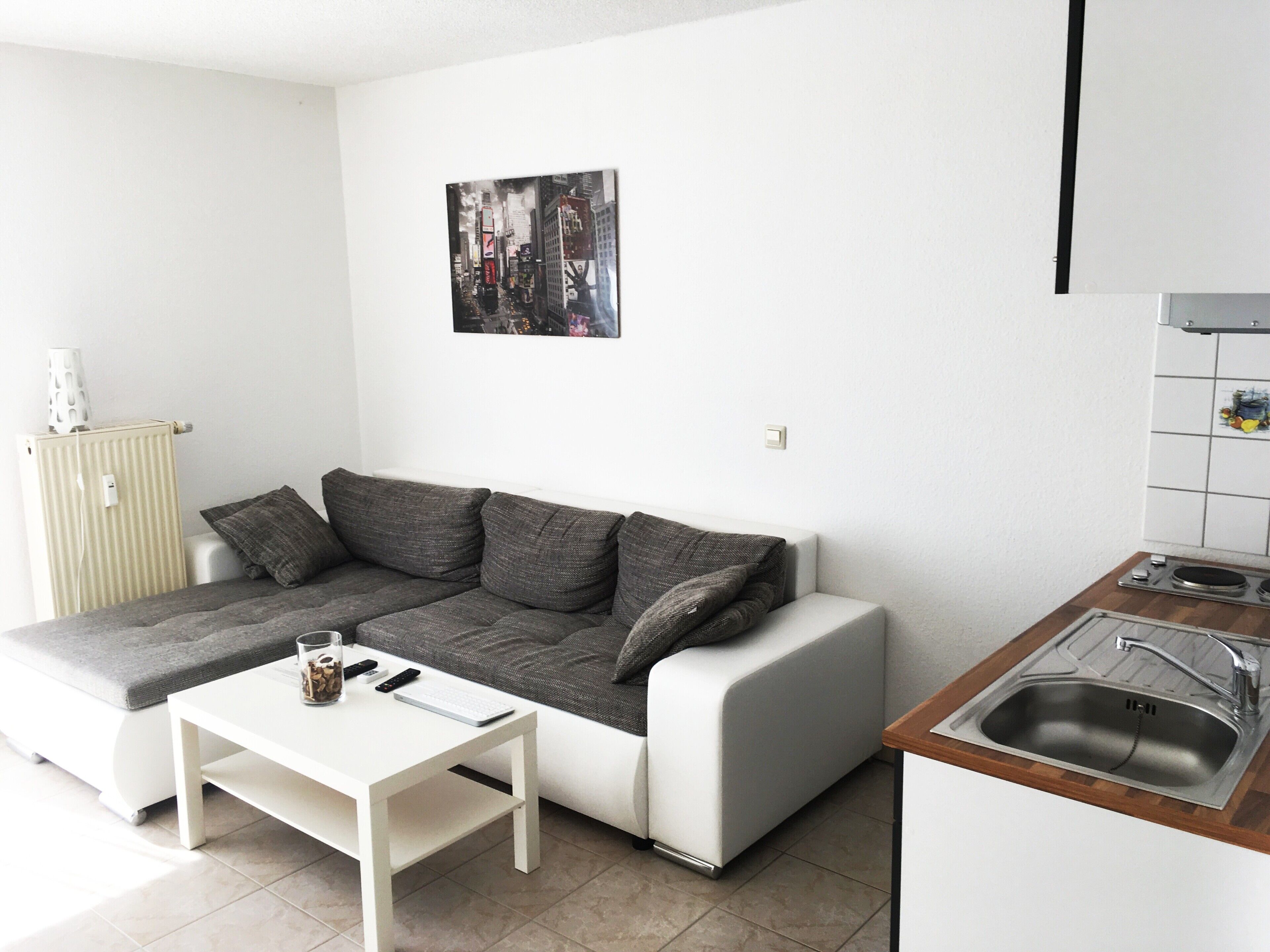 Un apartamento pequeño y moderno con sofá, una mesa blanca, una zona de cocina con fregadero y un radiador.